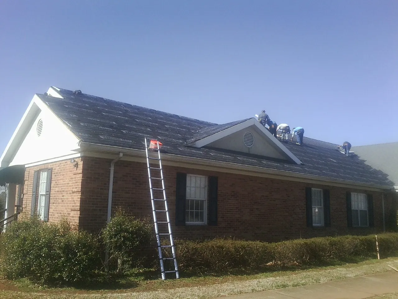 Crew tearing shingles off a brick ranch roof with a ladder set — Emergency Roof Repair work by our crew in Hiltons